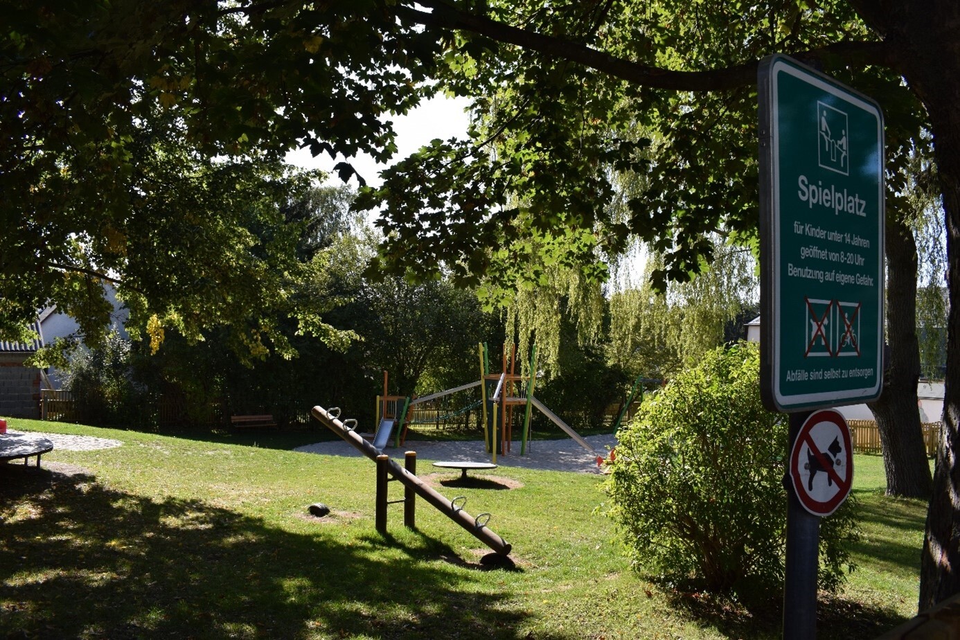 Eine Wippe im grünen auf einem idylischen Spielplatz. Von Bäumen umgeben. Im Vordergrund steht ein Schild mit dem Titel "Spielplatz" Im Hintergrund sind typische Spielgeräte zusehen.  (Link öffnet sich in einem Popupfenster mit vergrößerter Ansicht des Bildes.)