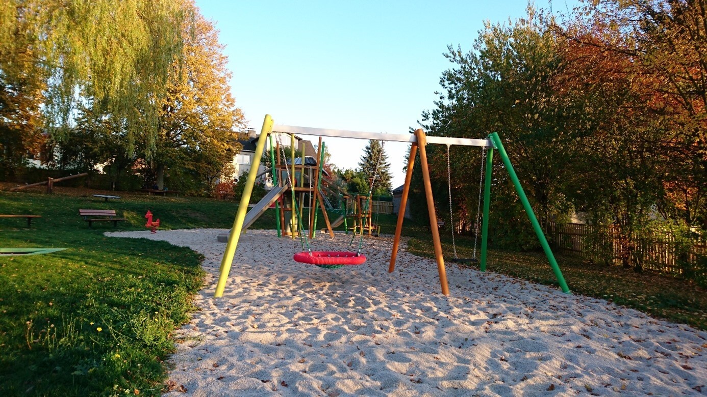 Ein buntes Schaukelgestell steht im Sand. Dahinter sind typische Spielplatzgeräte zu erkennen.  (Link öffnet sich in einem Popupfenster mit vergrößerter Ansicht des Bildes.)