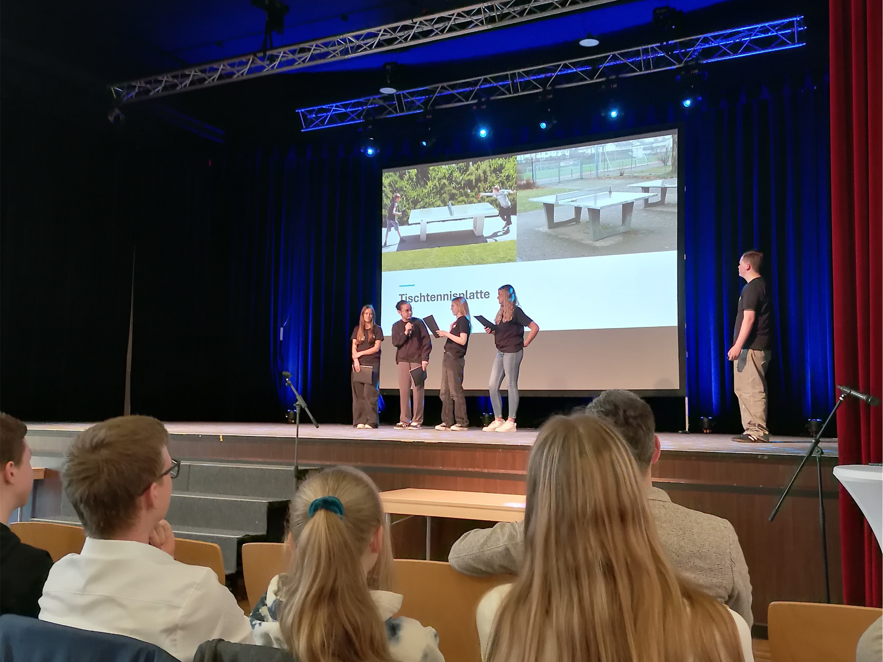 Auf dem Bild sieht man eine Bühne auf der fünf Personen stehen. Es sieht aus als wäre die rechte Person eine Art Moderator. Die weiteren vier Personen scheinen einen Vortrag zu halten. Hinter ihnen ist zudem eine Leinwand mit einem Bild zu sehen. Außerdem ist im Vordergrund des Bildes ein Teil des Publikums zu sehen.